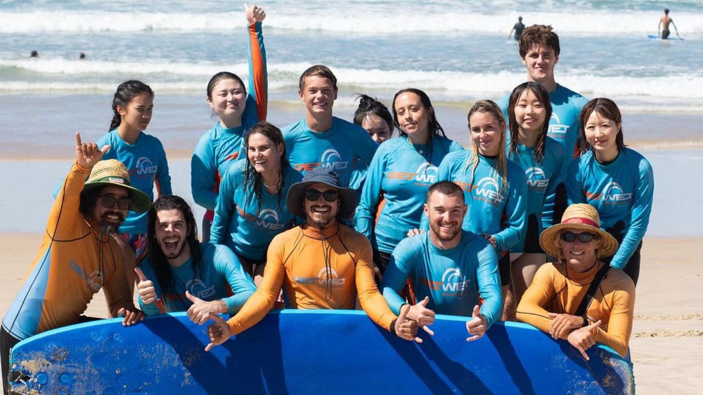 Budds hostel guest on the beach all standing behind a large surf board