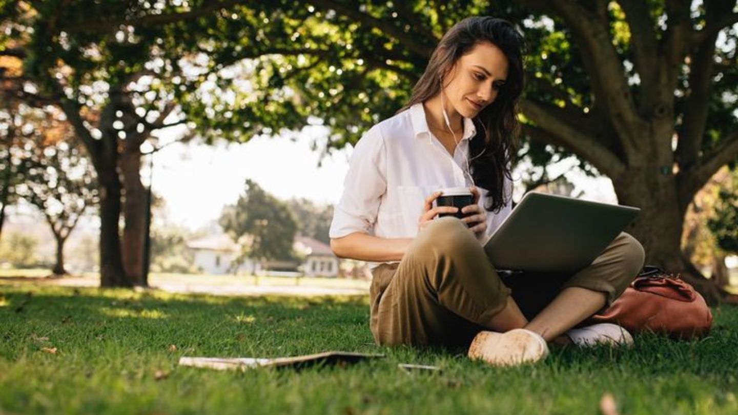 Backpacker with ear phones and sipping coffee whilst working on her laptop in a quiet park